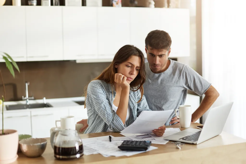 A young couple looking worried while reading financial documents in their kitchen.