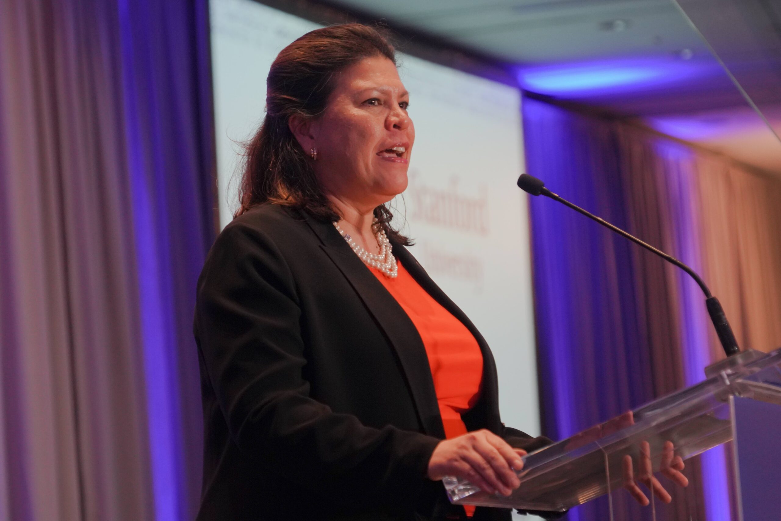 A woman in a black blazer and orange top speaks at a podium with a microphone, against a backdrop of purple-lit curtains and a projection screen.