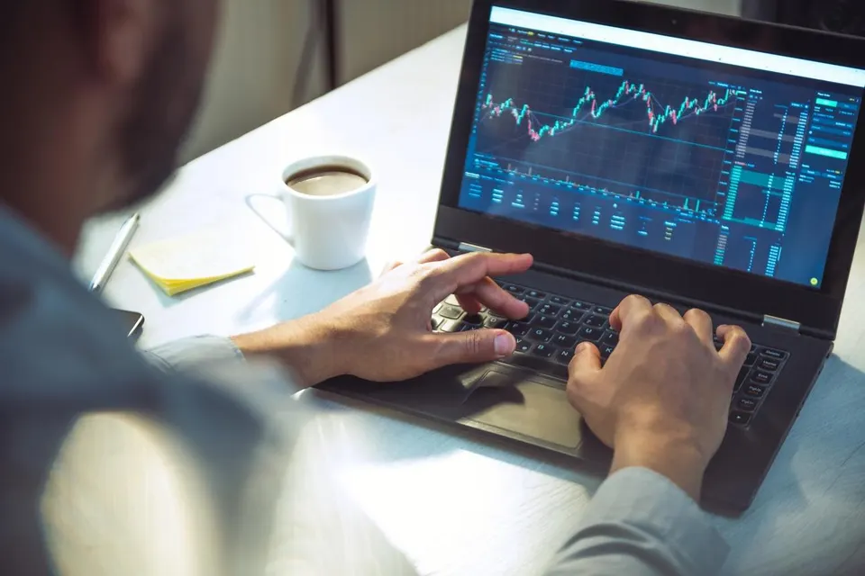 A young man is sits at a table with a laptop and a cup of coffee, checking stock market performance. 
