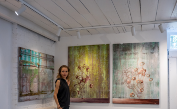 Miranda Donovan standing in her London studio beside three mixed-media paintings