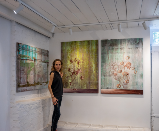 Miranda Donovan standing in her London studio beside three mixed-media paintings